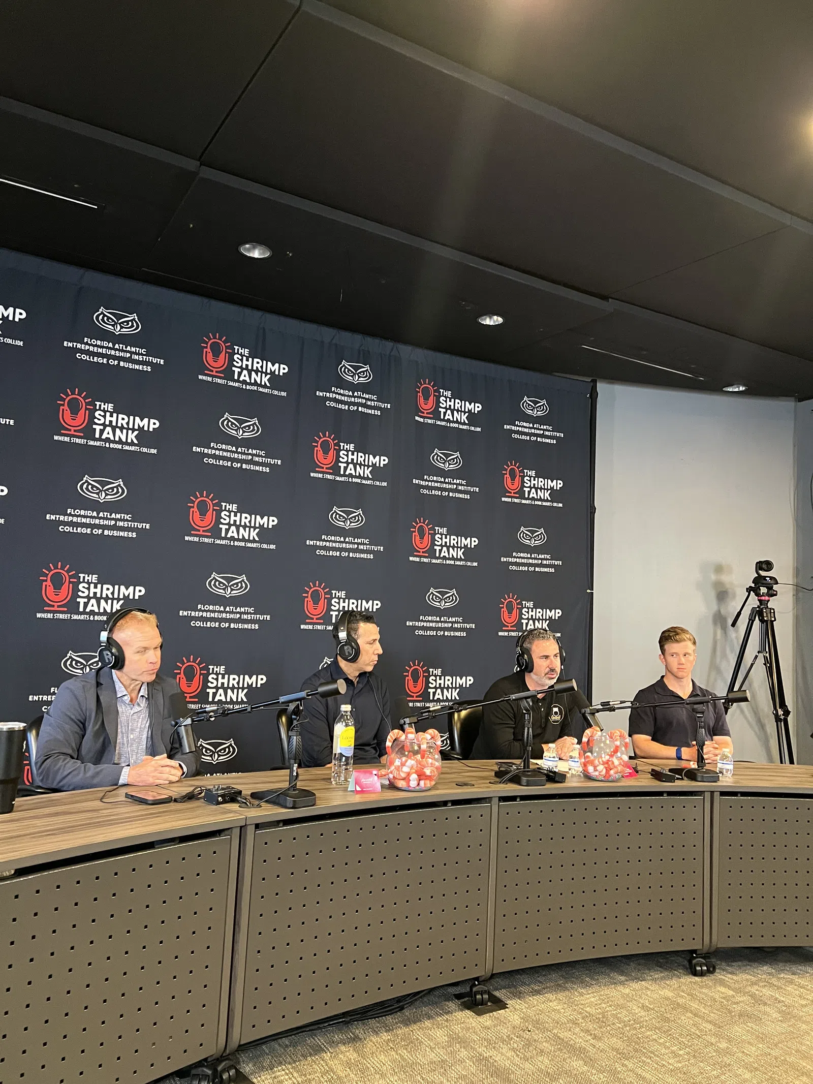 Four men wearing headsets sit behind microphones at a table in a podcast studio with The Shrimp Tank backdrop.