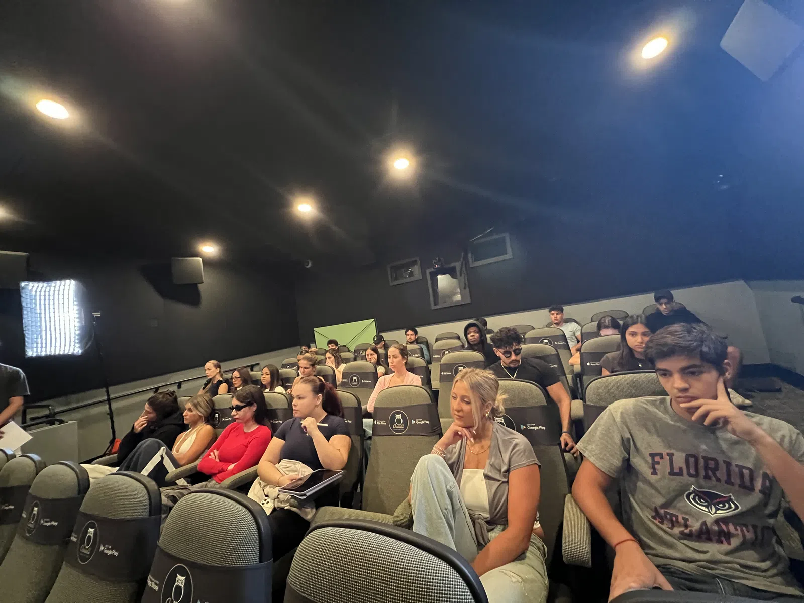 Students sitting attentively in a dimly lit lecture hall with padded seats and dark walls.