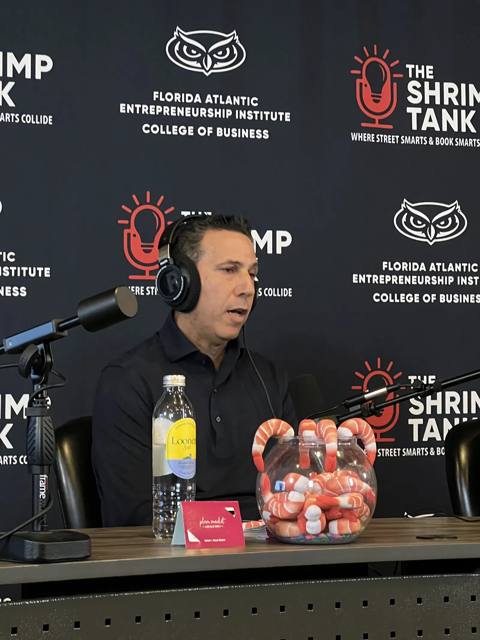 Man speaking into a microphone during a podcast at Florida Atlantic Entrepreneurship Institute with shrimp tank branding.