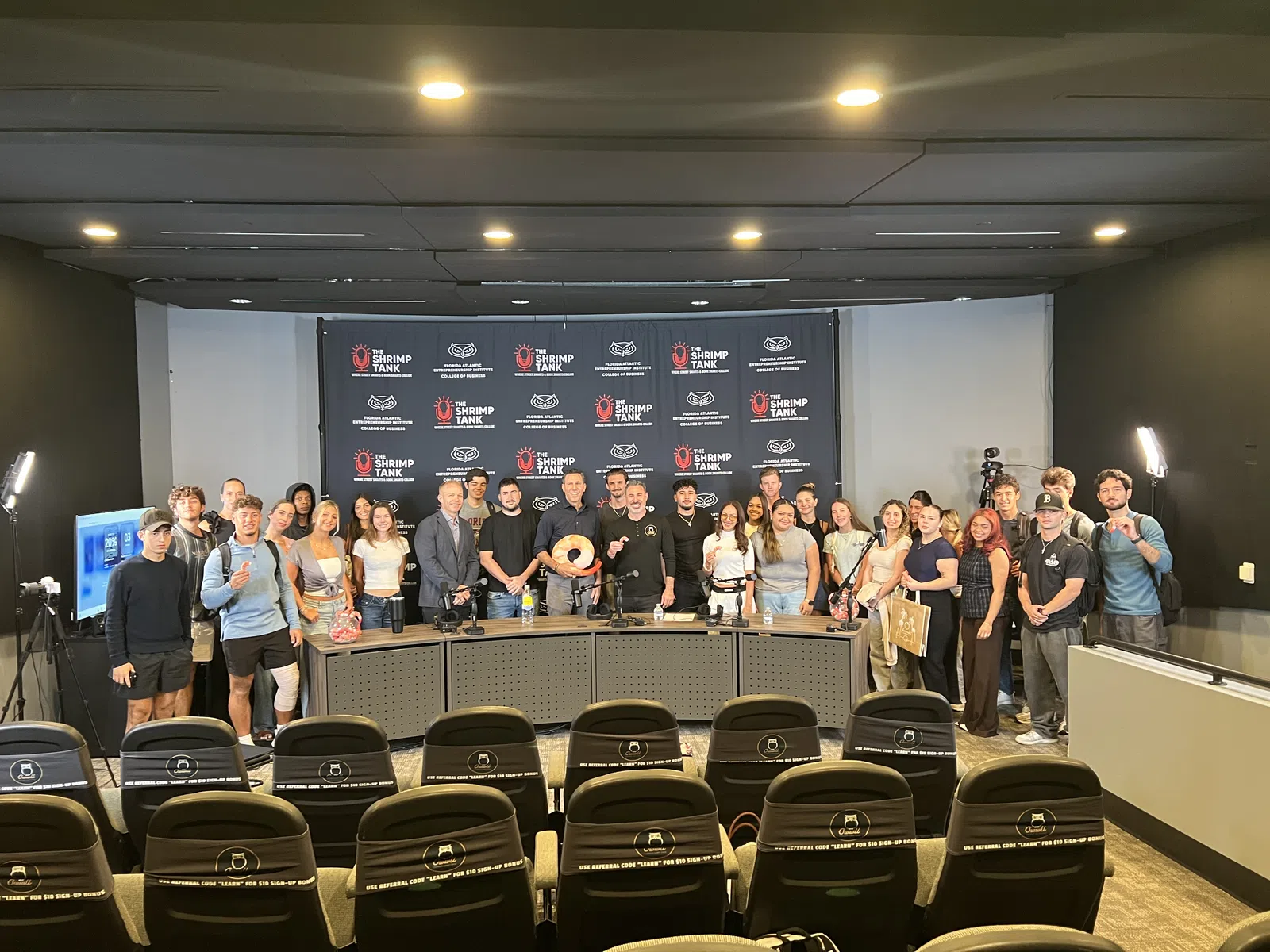 Group photo of diverse individuals standing in front of a Shrimp Tank event banner in a conference room.