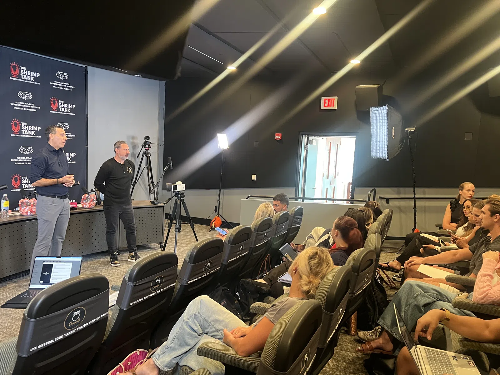 Two presenters speak to an audience in a small auditorium with cameras and lighting equipment set up.