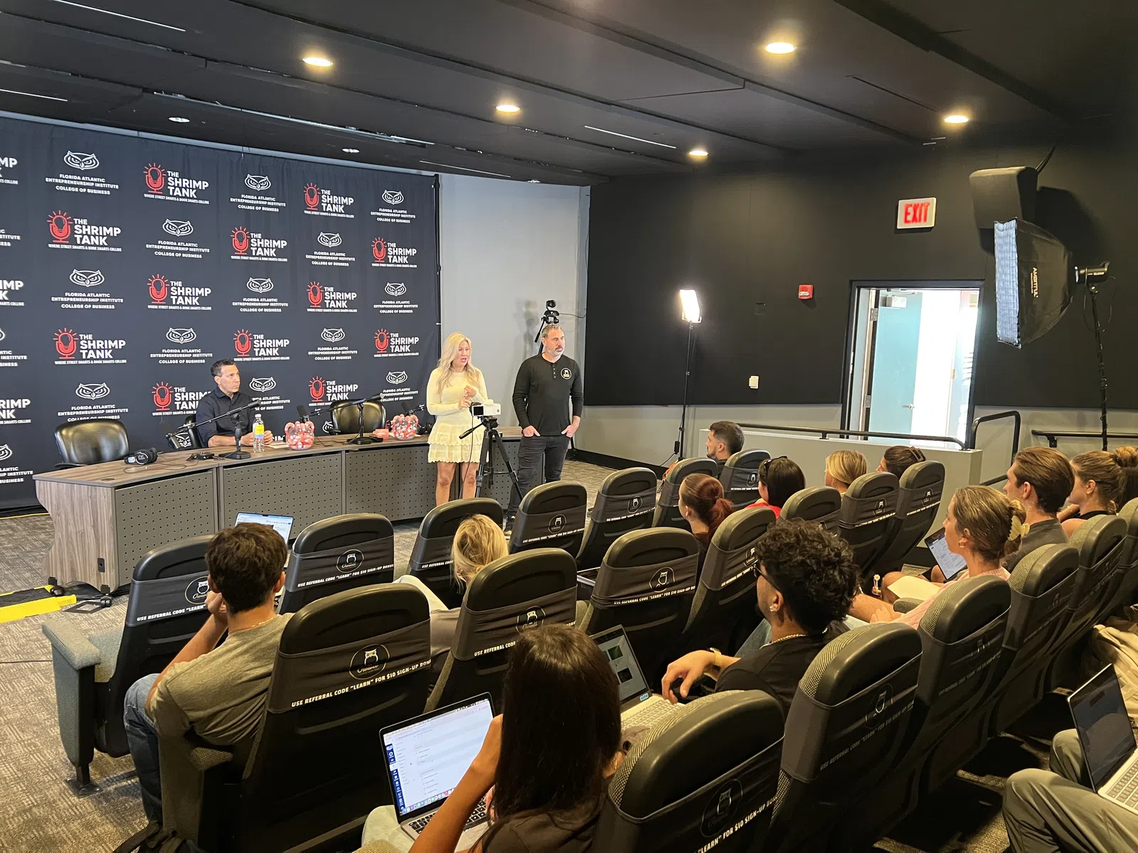 Presenters speaking to an attentive audience in a conference room with The Shrimp Tank branding on backdrop.