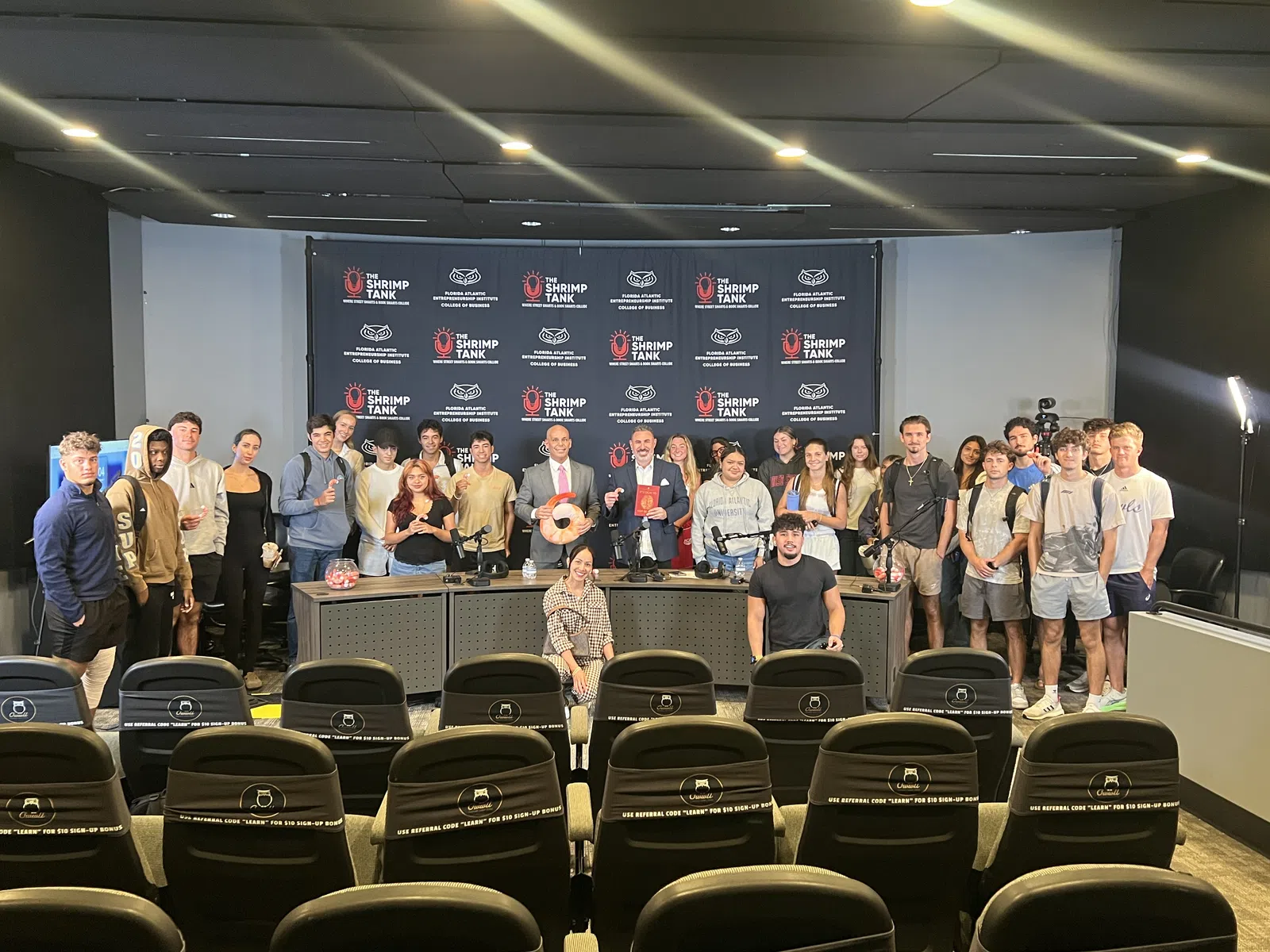 Group of young adults and hosts posing in a studio set for The Shrimp Tank with branded backdrop and chairs.