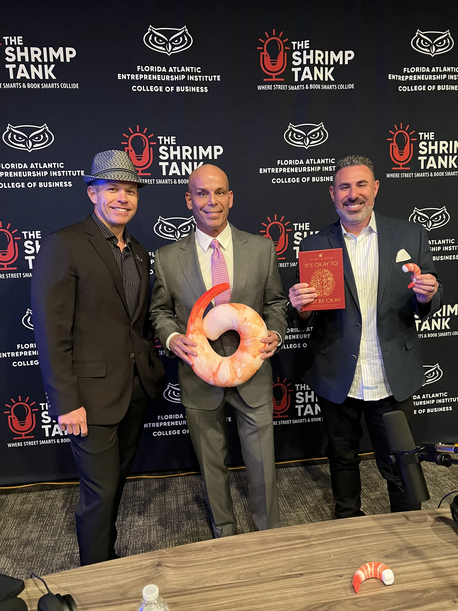 Three men in suits posing at The Shrimp Tank event with shrimp props and a book against branded backdrop.