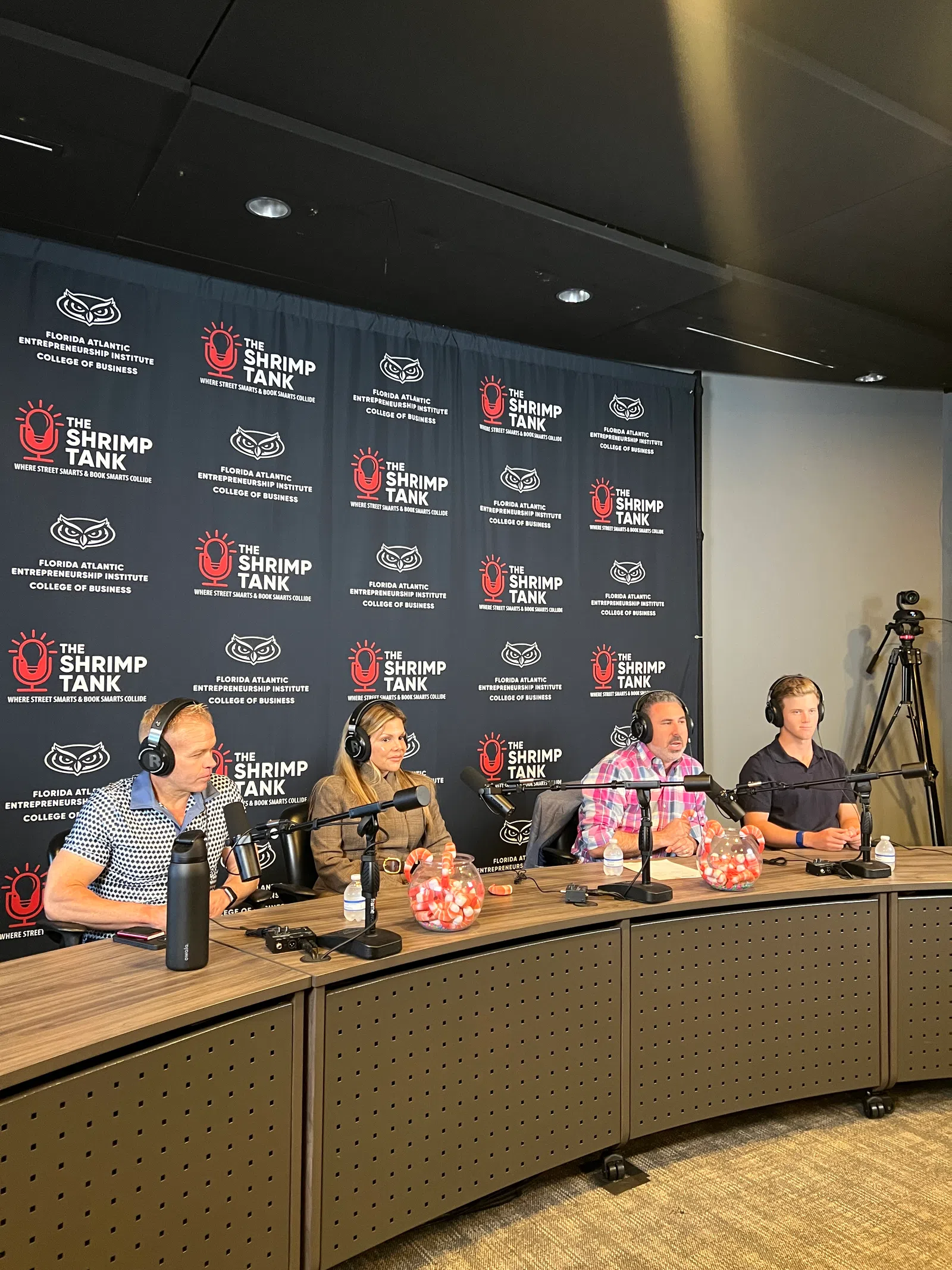 Four panelists with headphones sit at a podcast table discussing with microphones and branded backdrop for The Shrimp Tank.