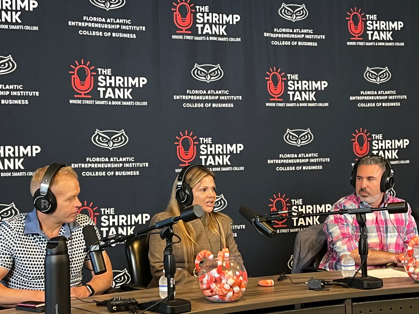 Three hosts wearing headphones speak into microphones during a podcast recording at The Shrimp Tank studio.