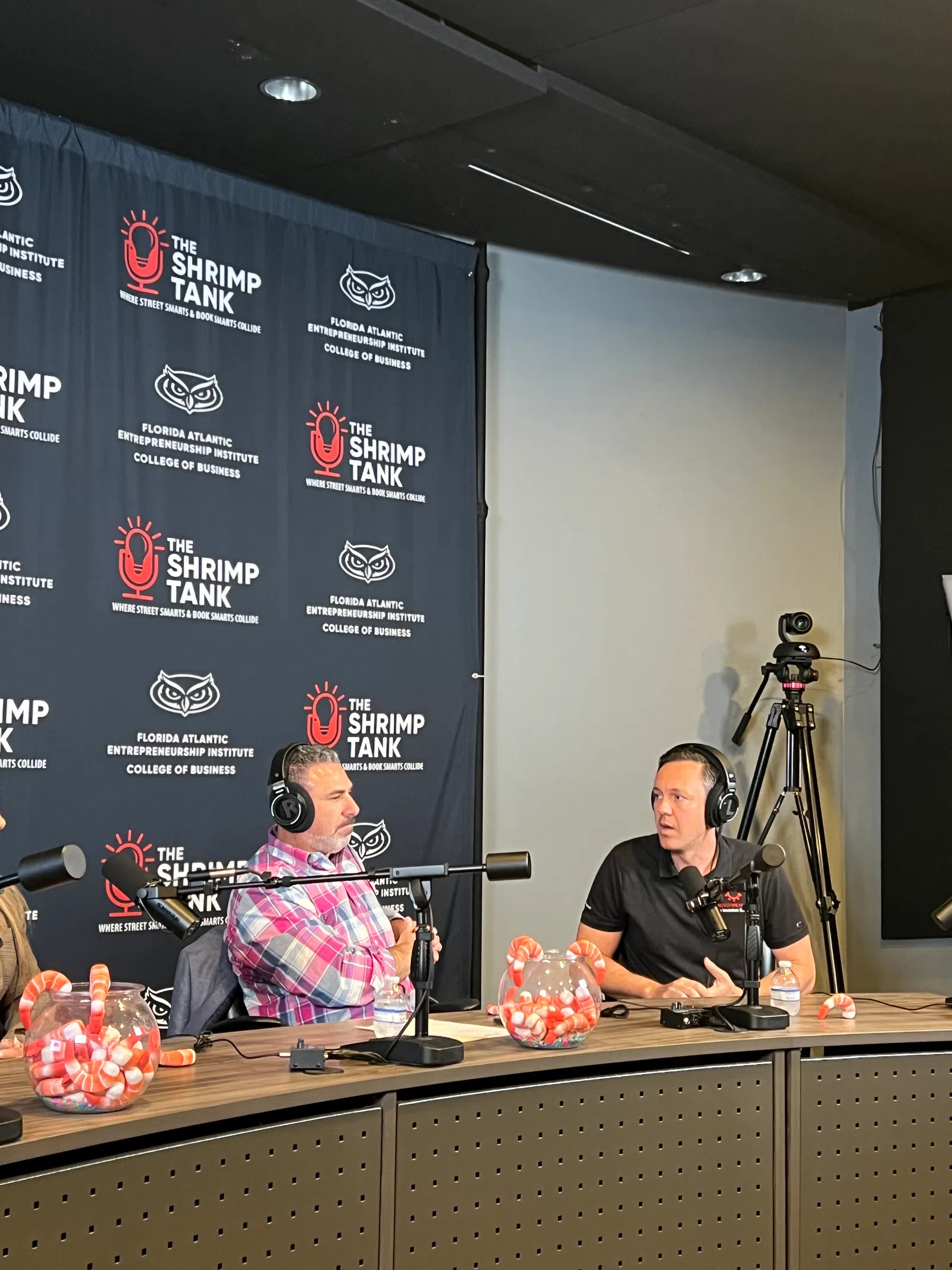 Two men wearing headphones discuss business on a podcast set with The Shrimp Tank backdrop and candy jars on the table.