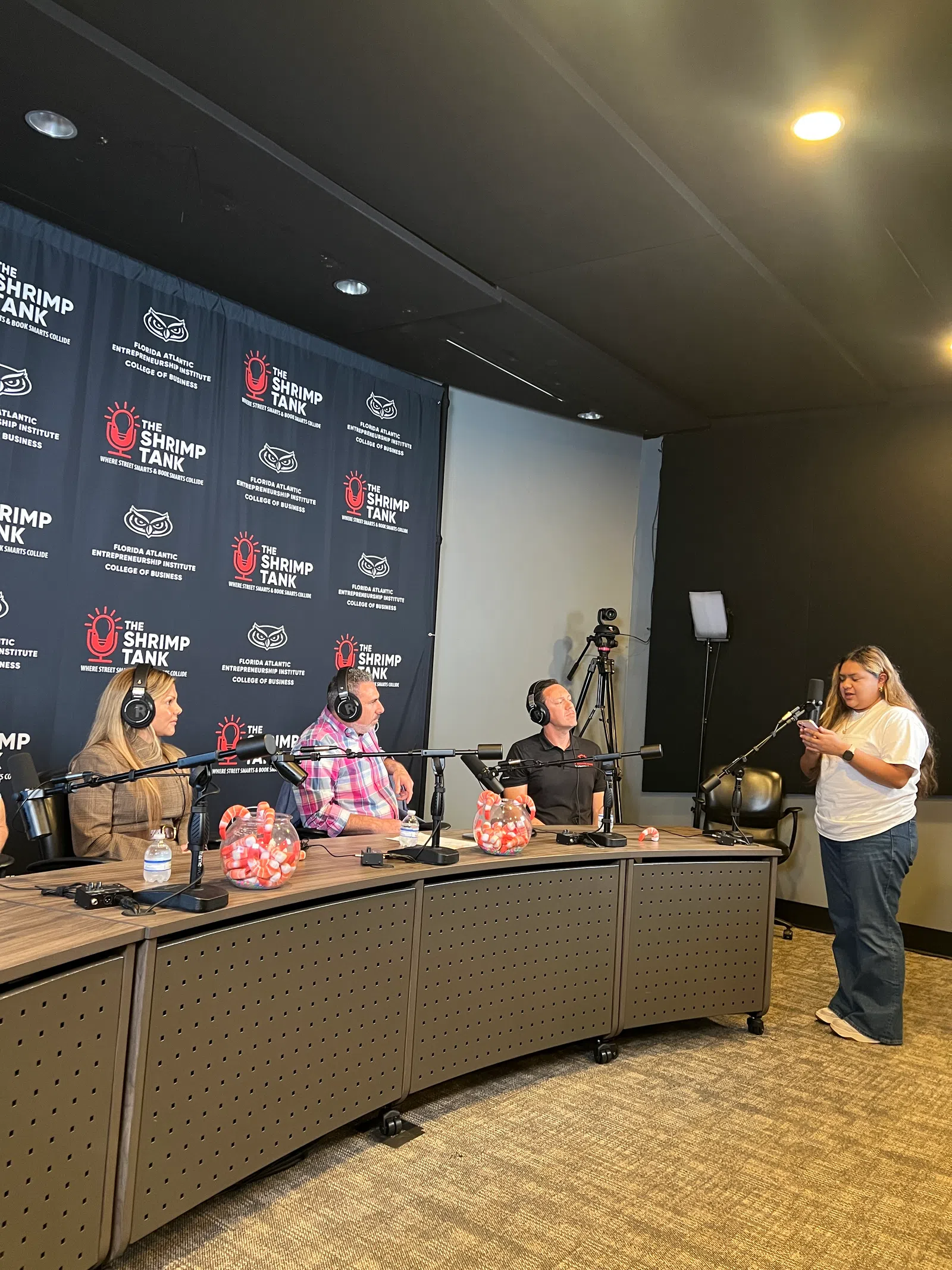 Three panelists wear headphones and speak into microphones at The Shrimp Tank event with a woman standing nearby.
