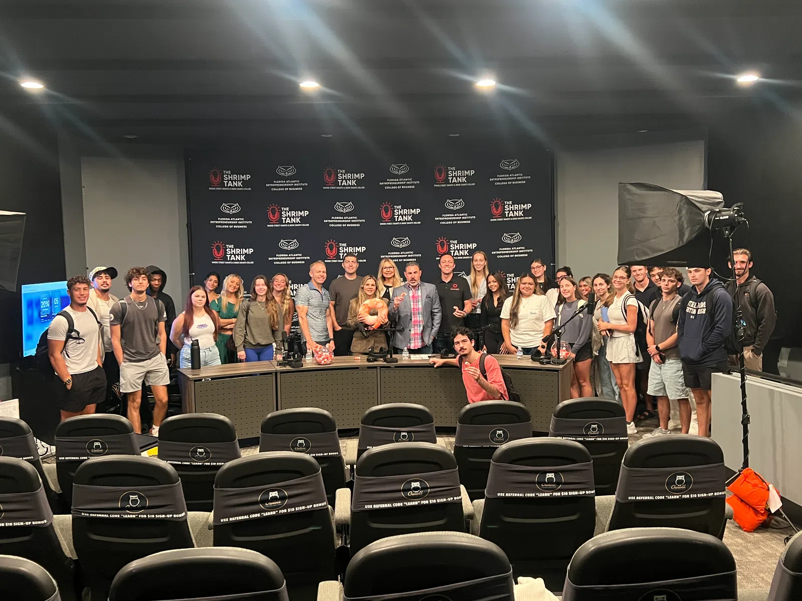 Group of people posing in a studio with The Shrimp Tank logos behind a panel and empty audience seats in front.