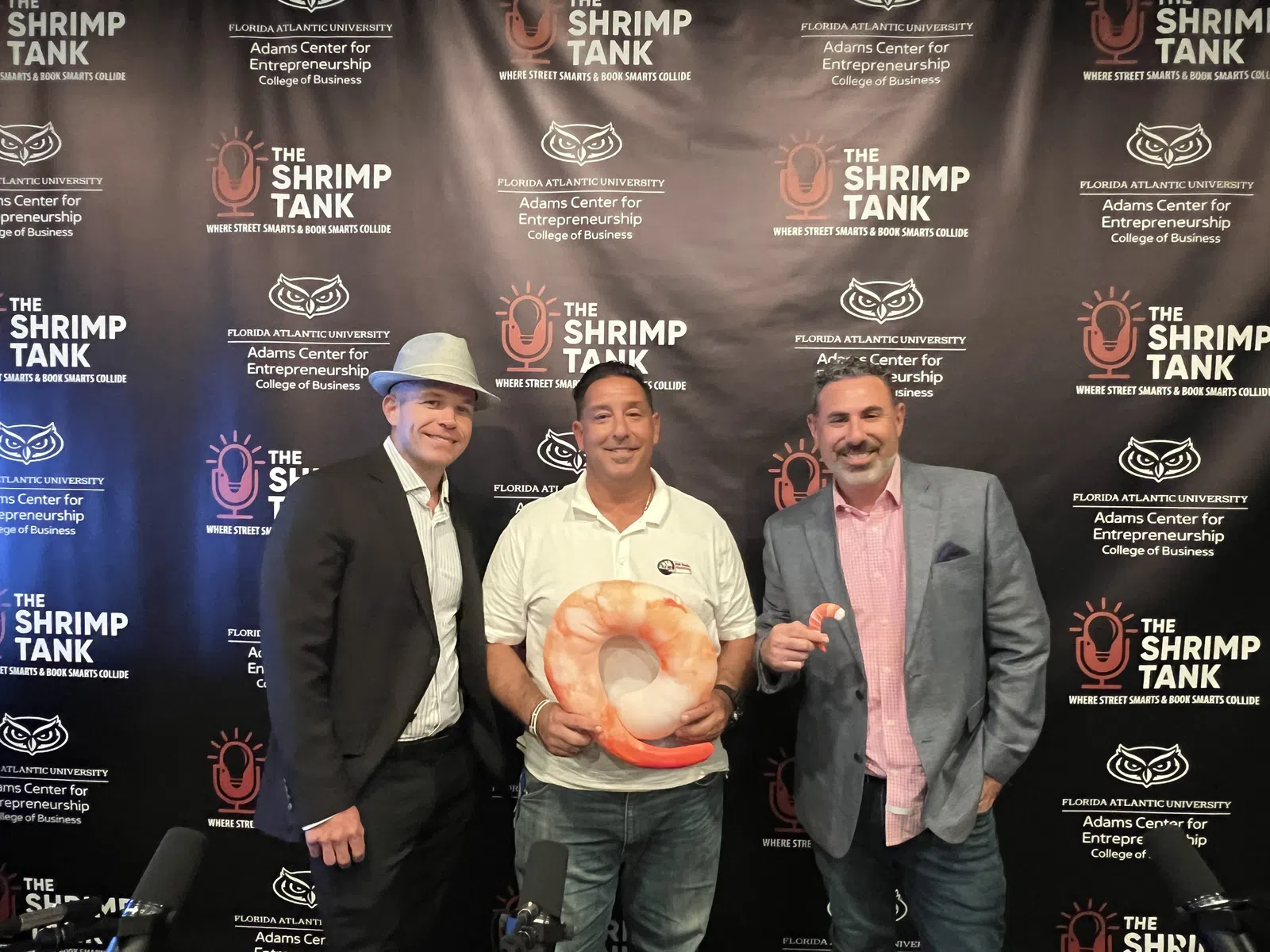 Three men posing in front of a Shrimp Tank backdrop, one holding a large shrimp prop, at an entrepreneurship event.