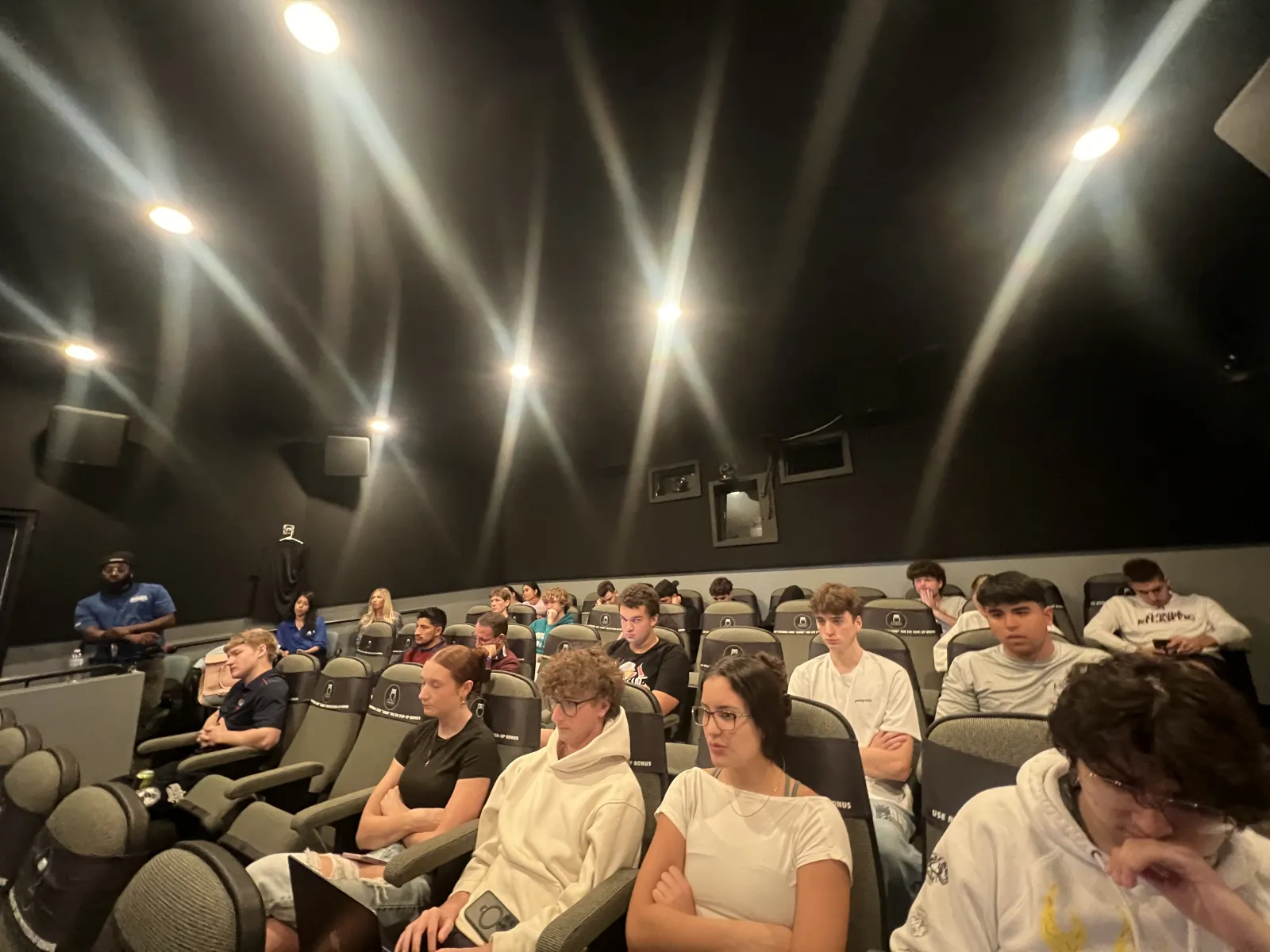 Group of young adults seated attentively in a dimly lit theater or lecture room with black walls and bright ceiling lights.