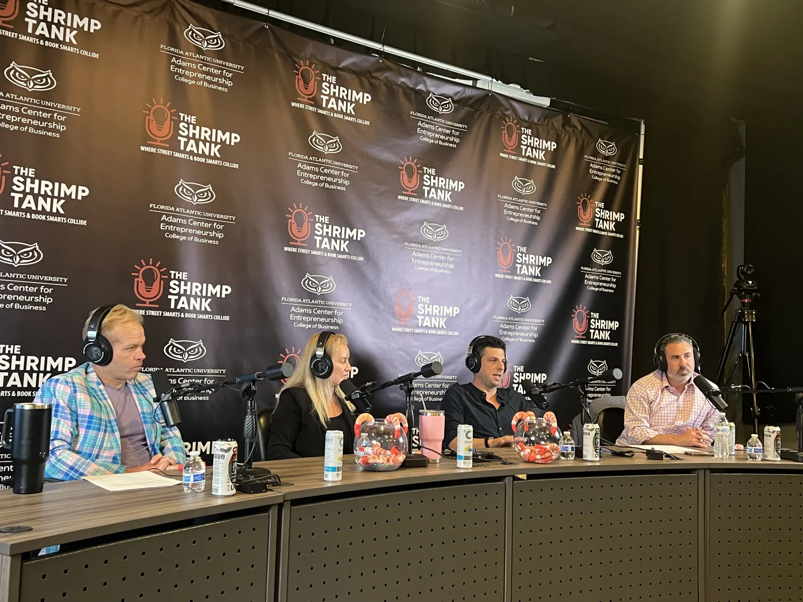 Four hosts with microphones and headphones recording a podcast at The Shrimp Tank event table with branded backdrop.