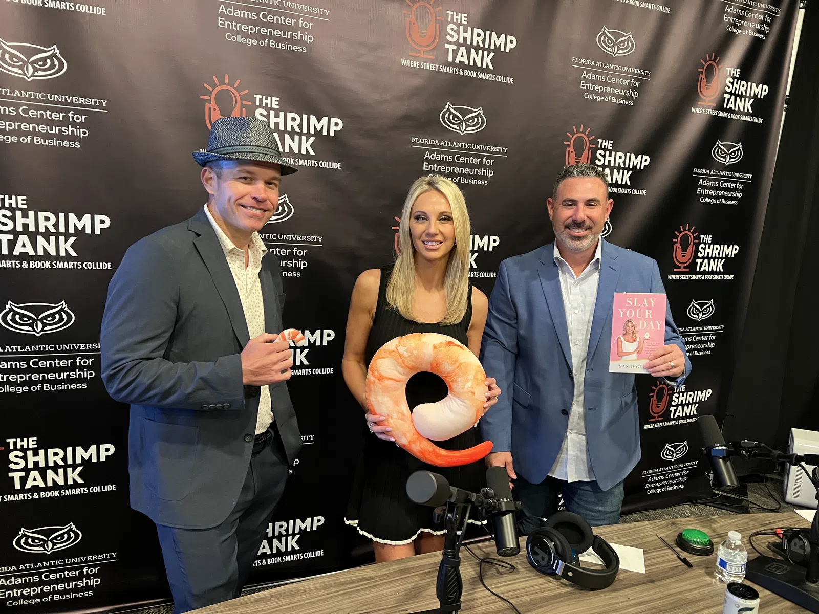 Three people posing with a shrimp-shaped pillow and a book in front of The Shrimp Tank event backdrop at Florida Atlantic University.