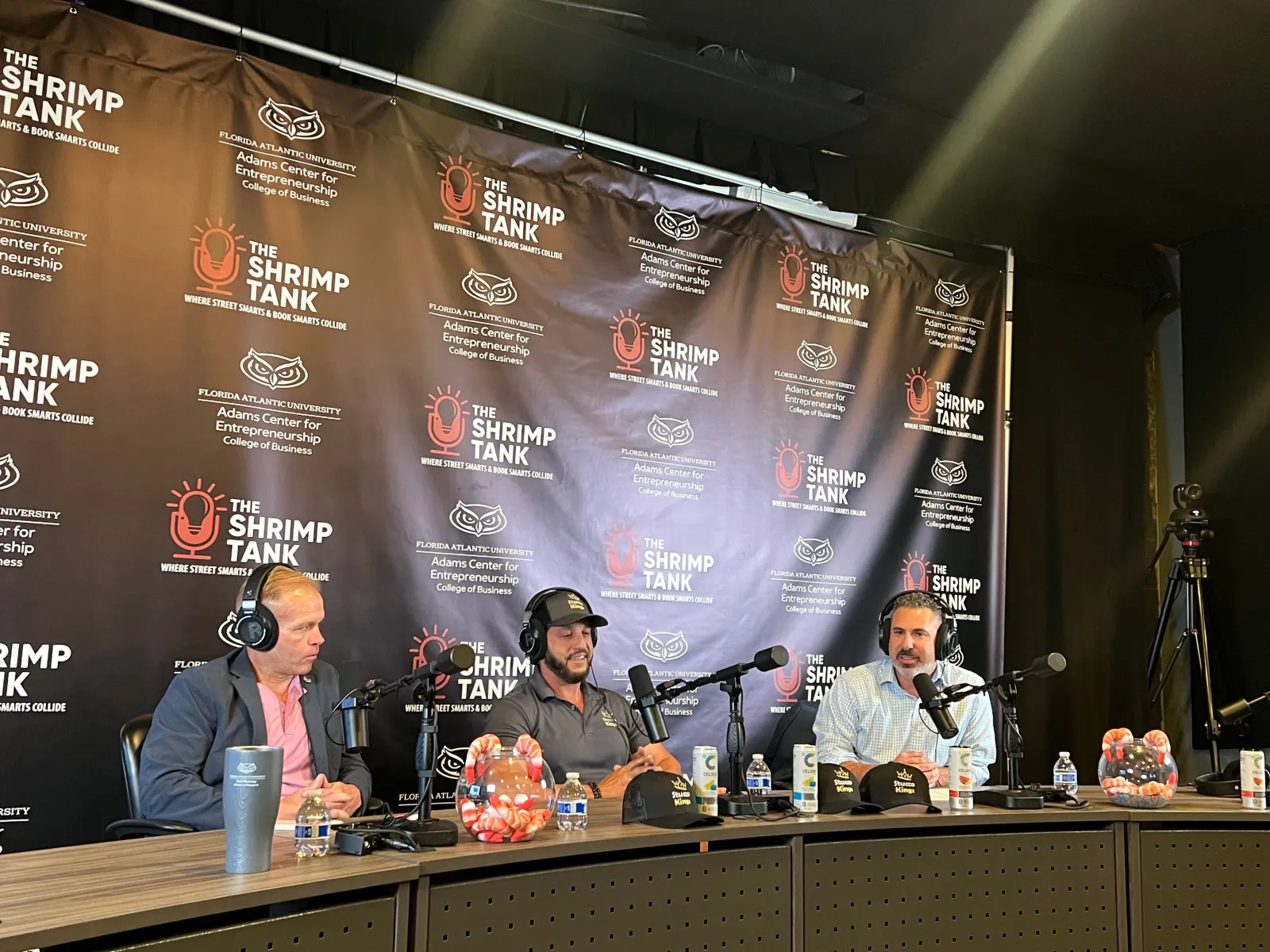 Three men with headsets and microphones seated at a table with The Shrimp Tank branded backdrop during a podcast or interview.