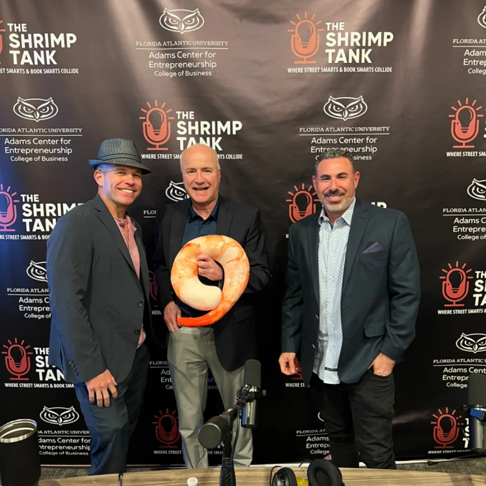 Three men posing at The Shrimp Tank entrepreneurship event with shrimp-shaped trophy and branded backdrop