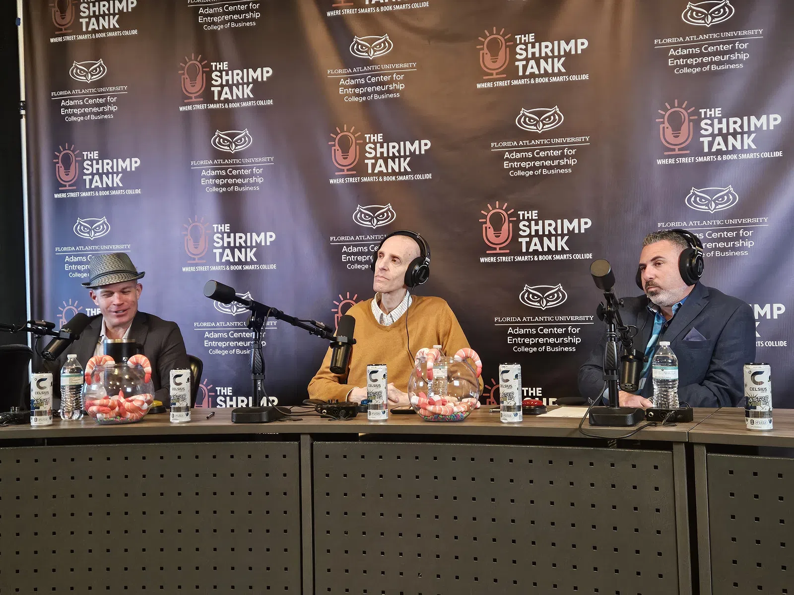 Three men with headphones sitting at a table with microphones and candy jars during The Shrimp Tank event backdrop.