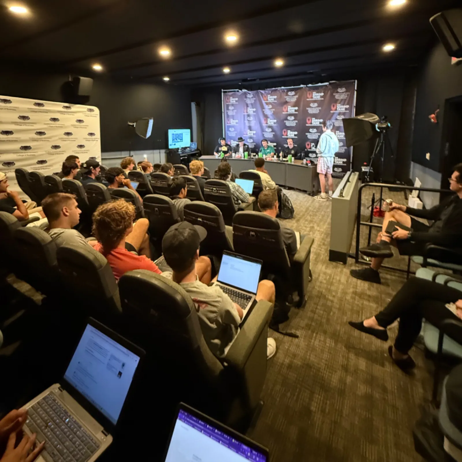 Audience attending a podcast panel discussion in a small theater room with laptops and microphones setup