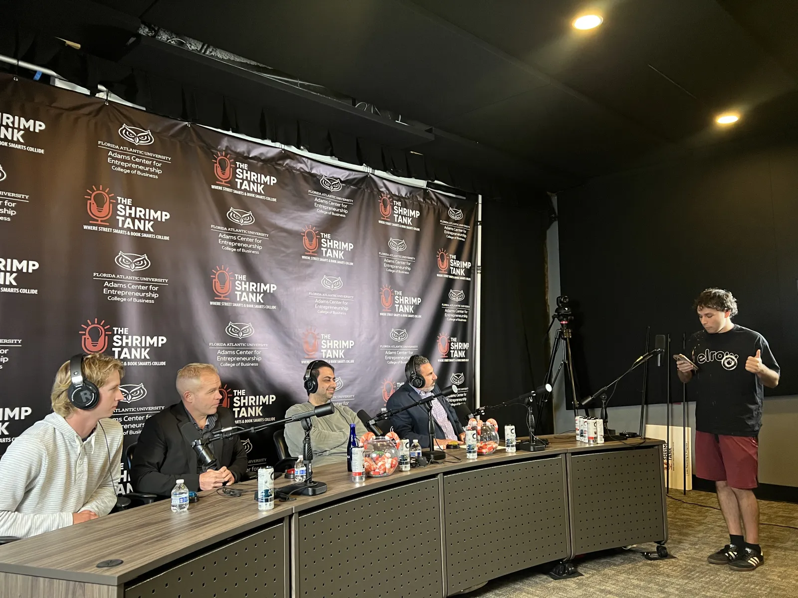 Four judges seated at a table with microphones listening to a standing contestant during The Shrimp Tank pitch event.