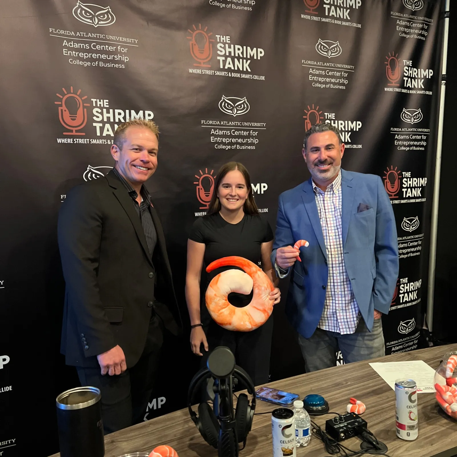 Three people posing at a Shrimp Tank event backdrop, one holding a large shrimp plush, with drinks and microphones nearby.