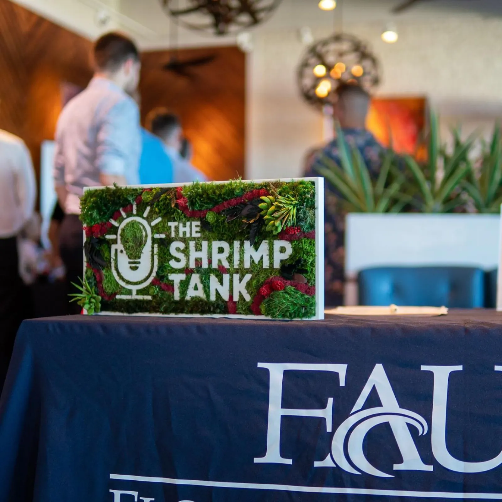 Decorative sign for The Shrimp Tank event on a table with FAU branding and blurred people in the background