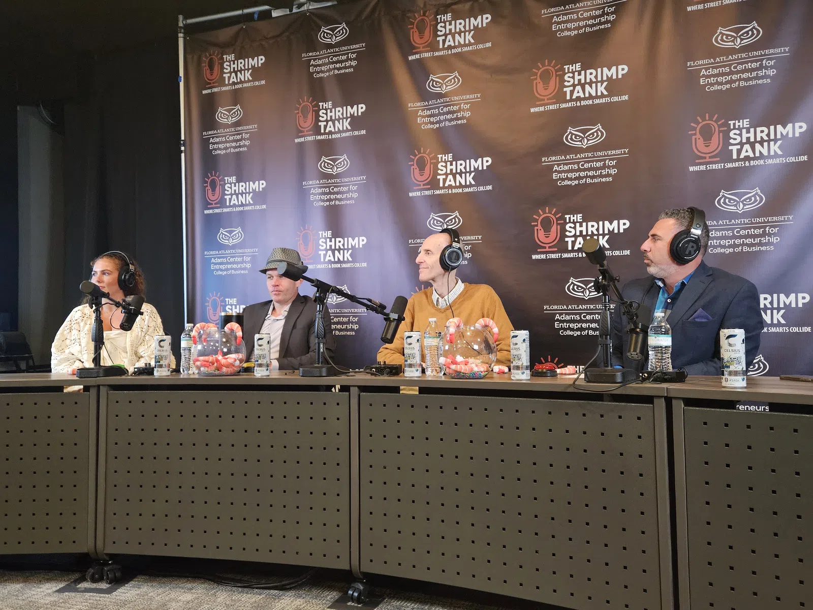 Four podcast hosts wearing headphones and speaking into microphones at The Shrimp Tank event panel.