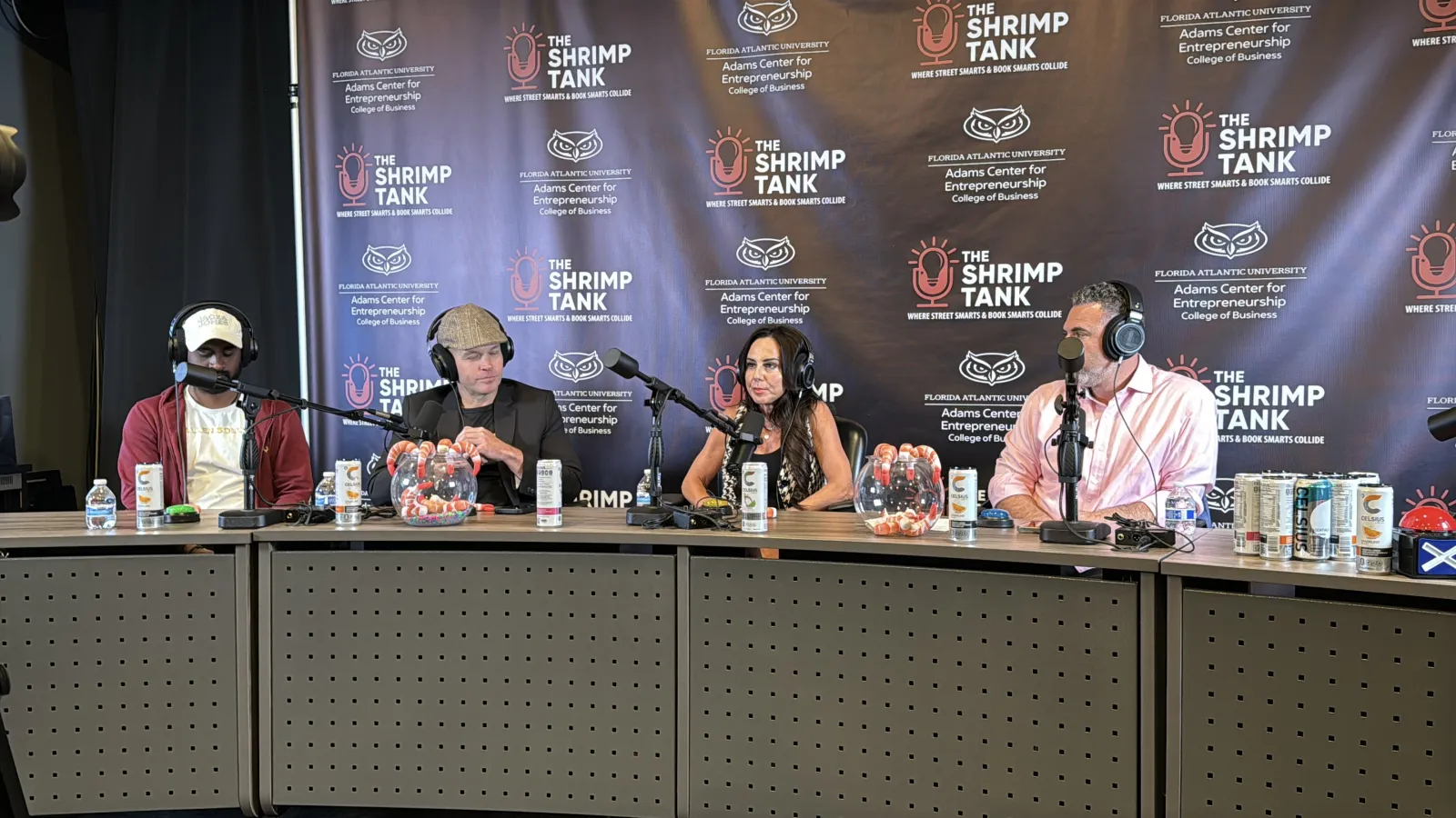 Four hosts wearing headphones speak into microphones during The Shrimp Tank podcast recording with branded backdrop.