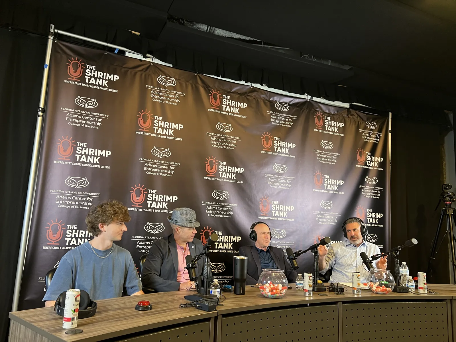 Four men seated at a podcast table with microphones and drinks against a The Shrimp Tank branded backdrop.