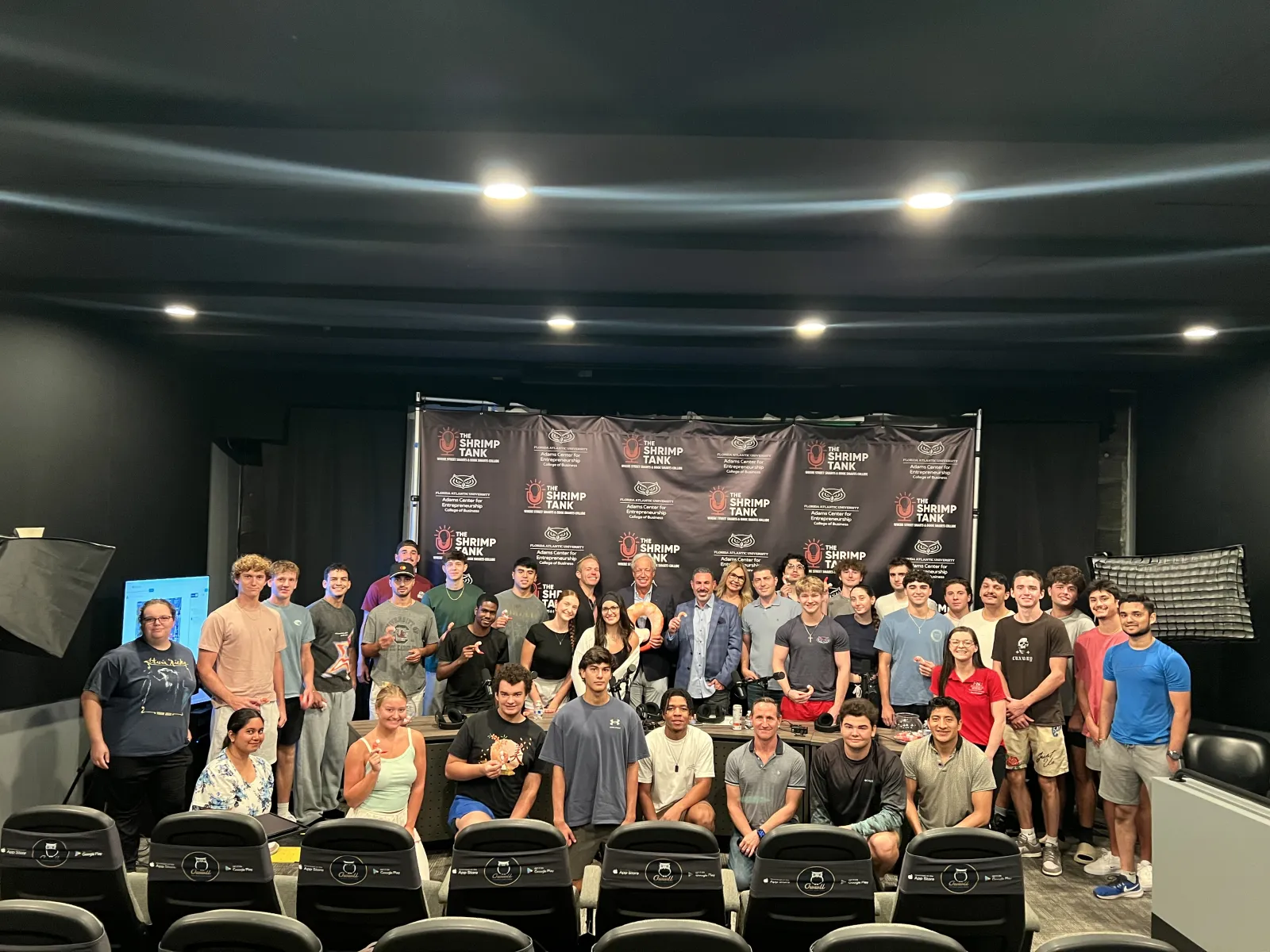 Large group of diverse young adults posing in front of Shrimp Tank banner in well-lit indoor event space.