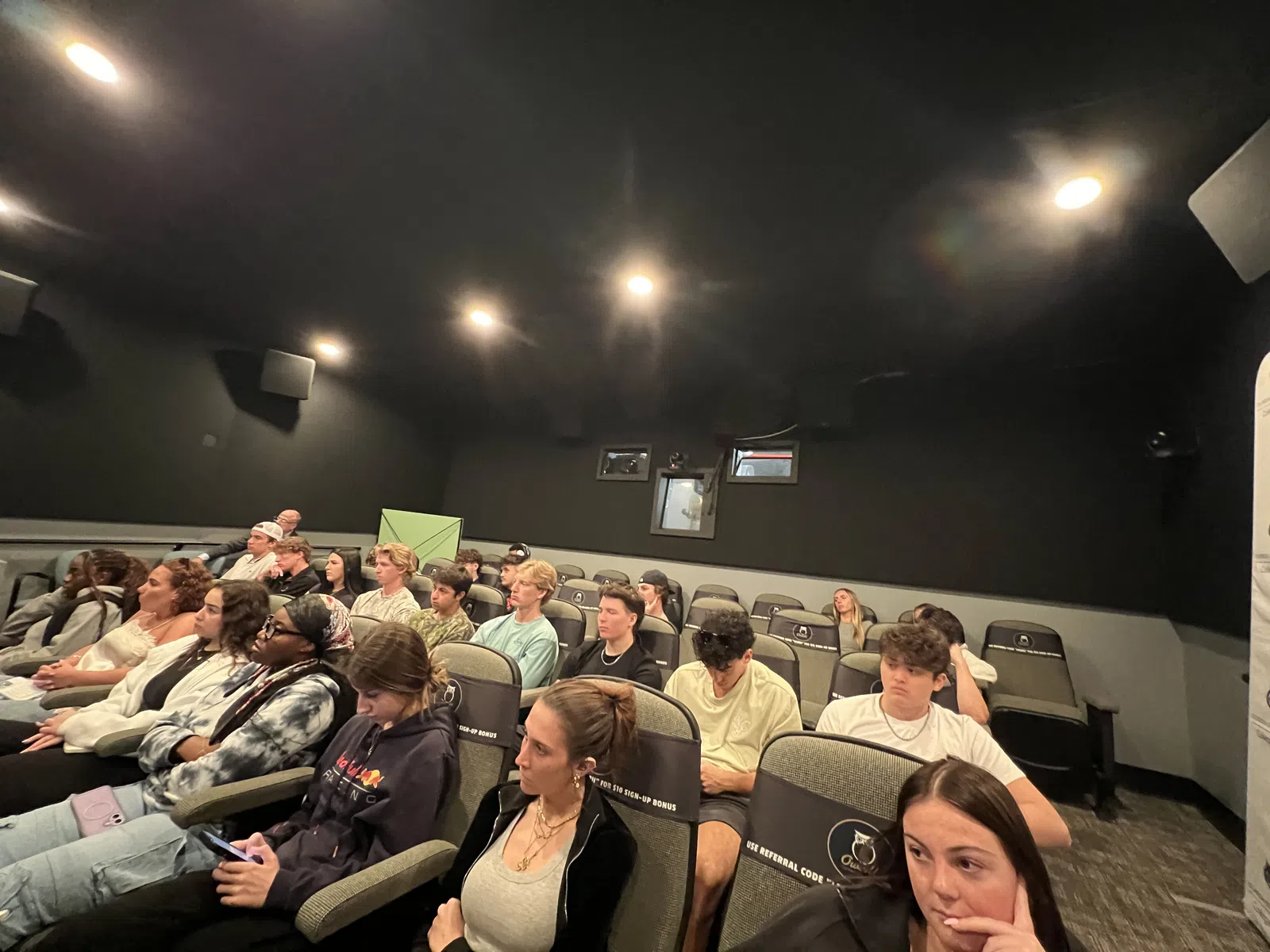 A diverse group of young adults attentively watching a presentation in a dimly lit auditorium with black walls.