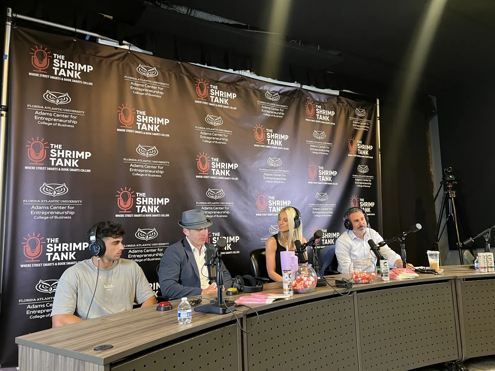 Four podcast hosts with microphones seated at a table during The Shrimp Tank by Florida Atlantic University event.