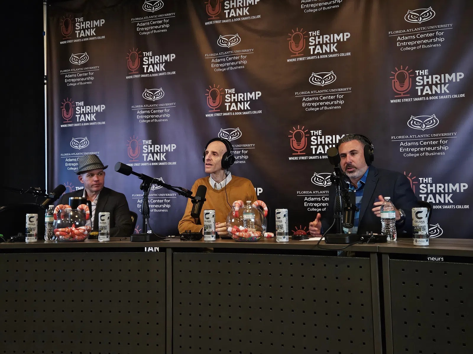 Three men seated at a podcast table with microphones and branding backdrop for The Shrimp Tank event
