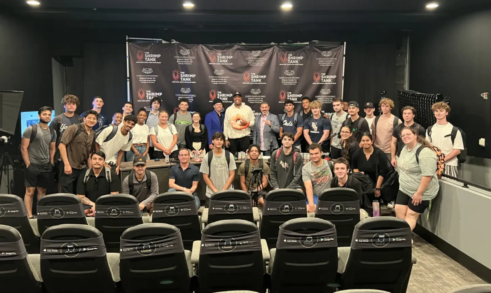 Group of young adults and instructors posing for a photo in a studio with Shrimp Tank logos on the background.