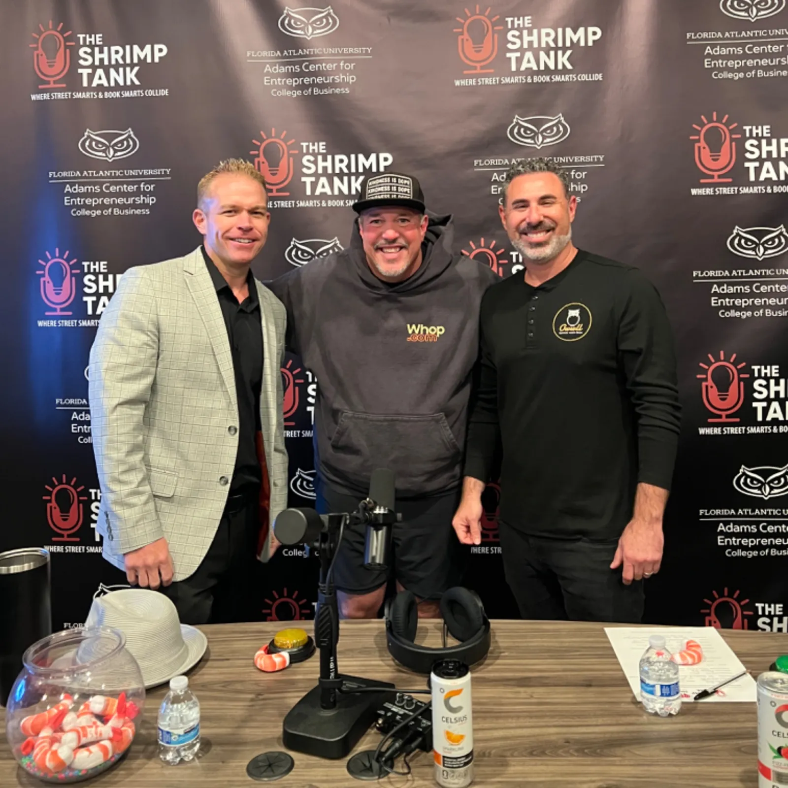 Three men posing in front of The Shrimp Tank podcast backdrop at a recording table with microphones and refreshments.