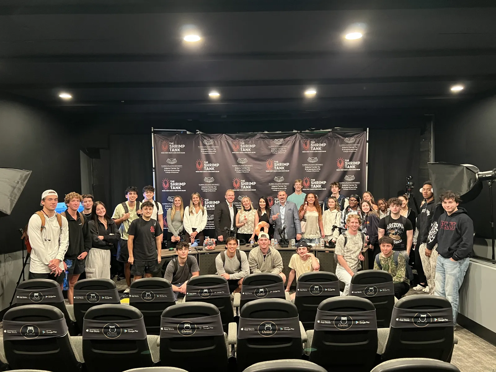 Group photo of diverse young people and adults posing in front of a branded backdrop in a studio setting.
