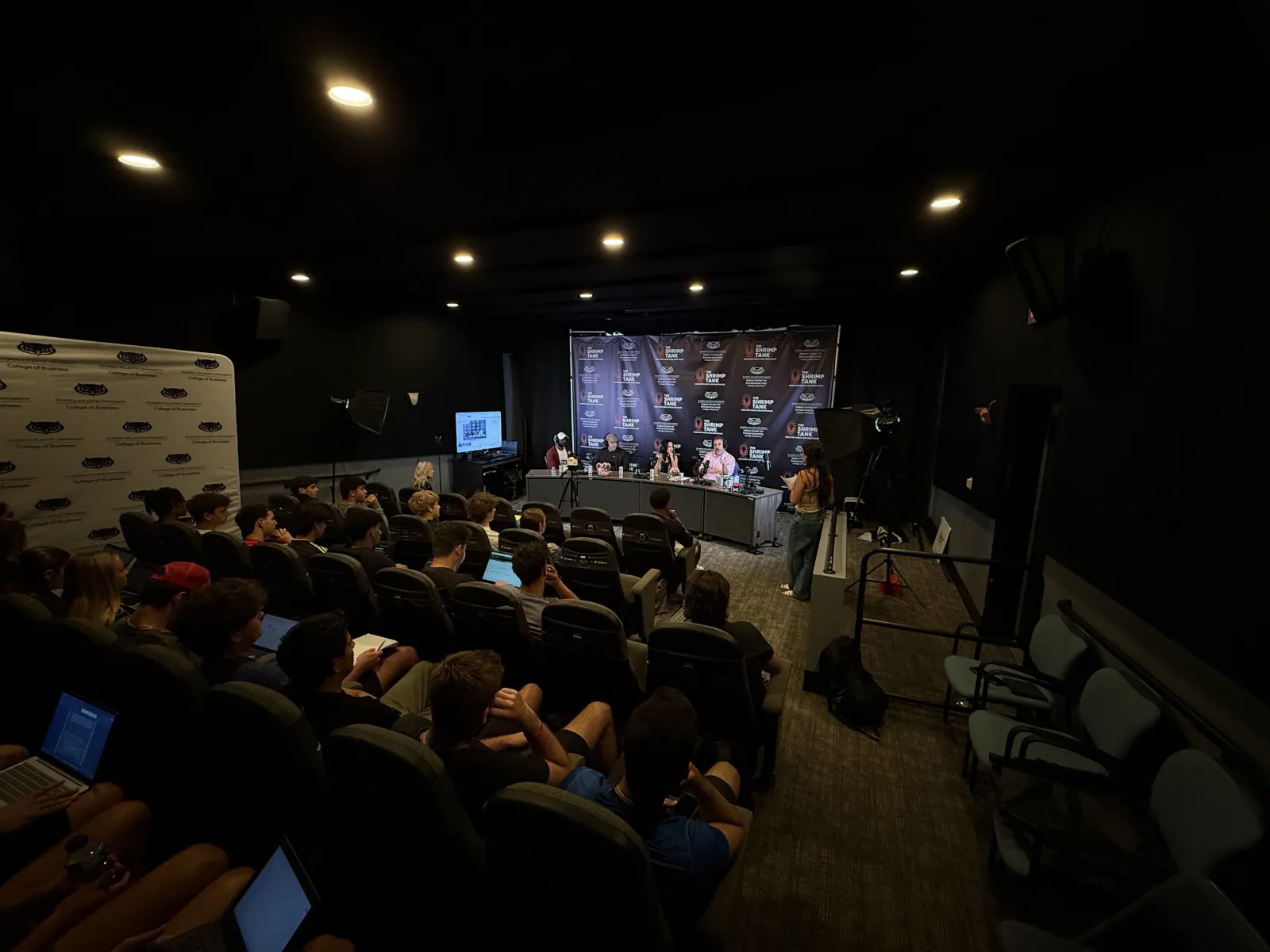 Audience attentively watching a panel discussion in a dark conference room with speakers at a table and media equipment.