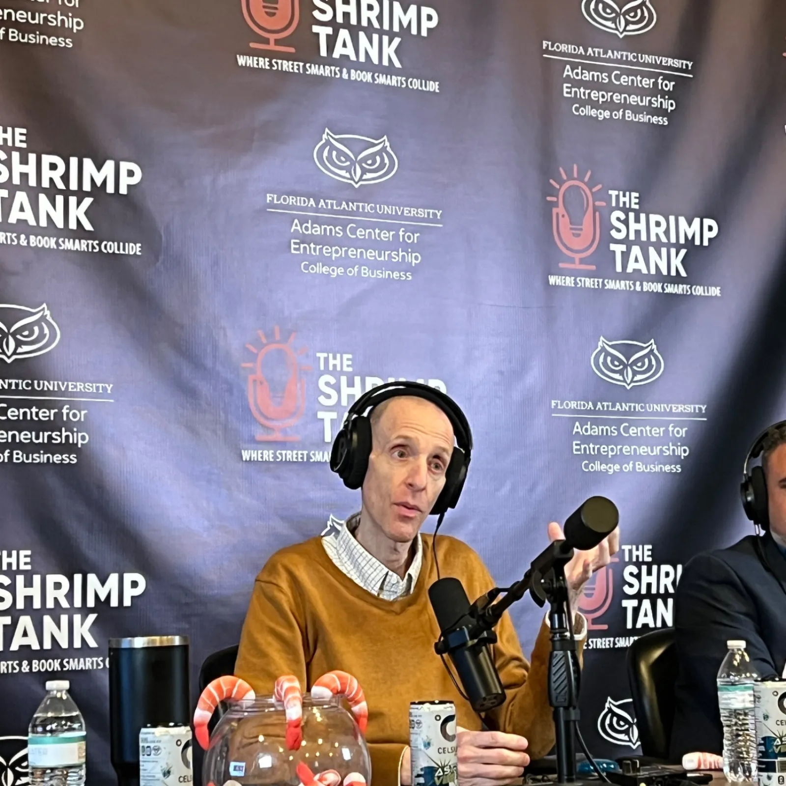 Man wearing headphones speaking into a microphone at The Shrimp Tank podcast with branded backdrop and shrimp jar.