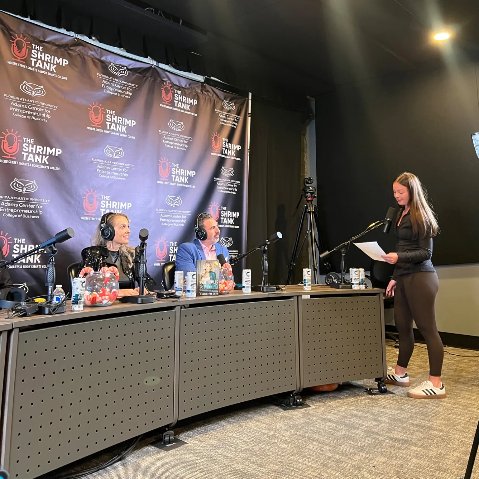 Radio hosts in headphones recording a podcast with a standing woman speaking into a microphone in a studio.