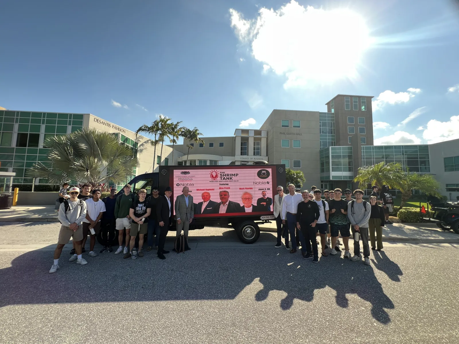 Group of people posing outdoors in front of a trailer with Shrimp Tank display at a sunny campus with palm trees.
