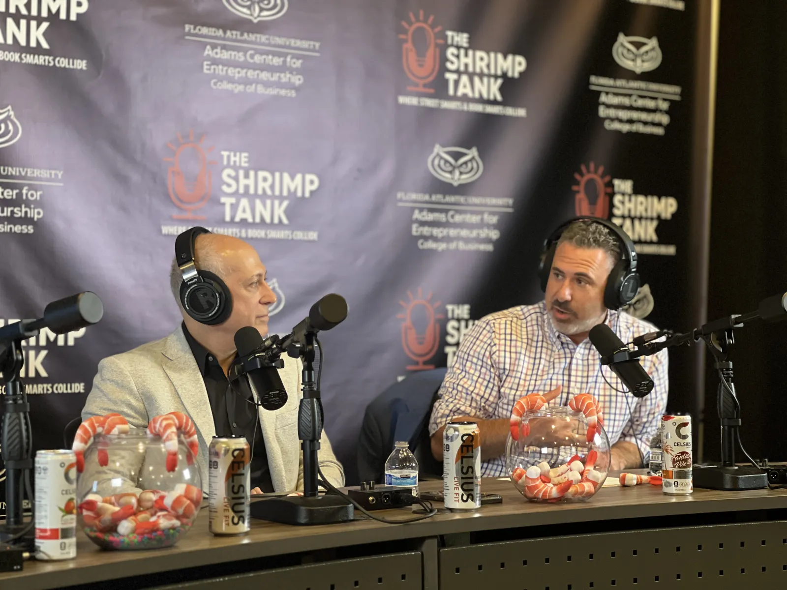Two men wearing headphones discussing on a podcast set with shrimp and Celsius drink cans on the table