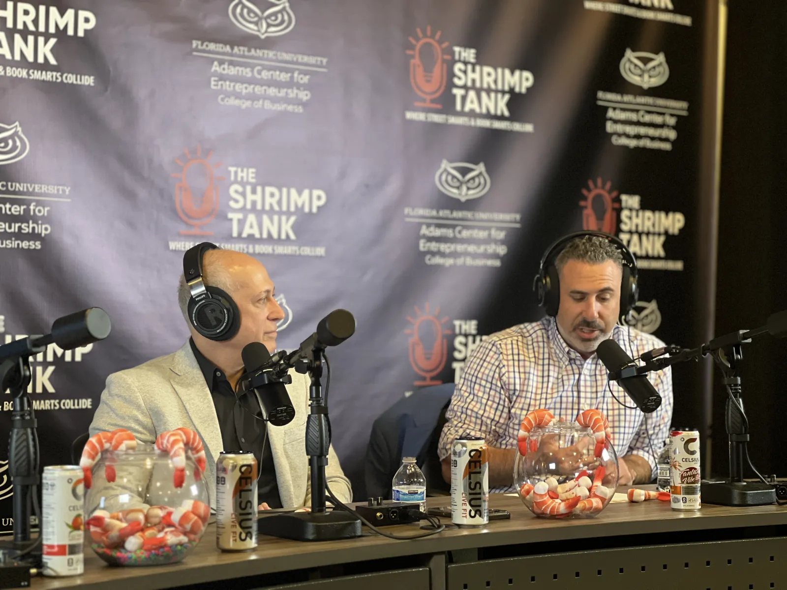 Two men with headphones speaking into microphones at The Shrimp Tank podcast with shrimp and soda on the table