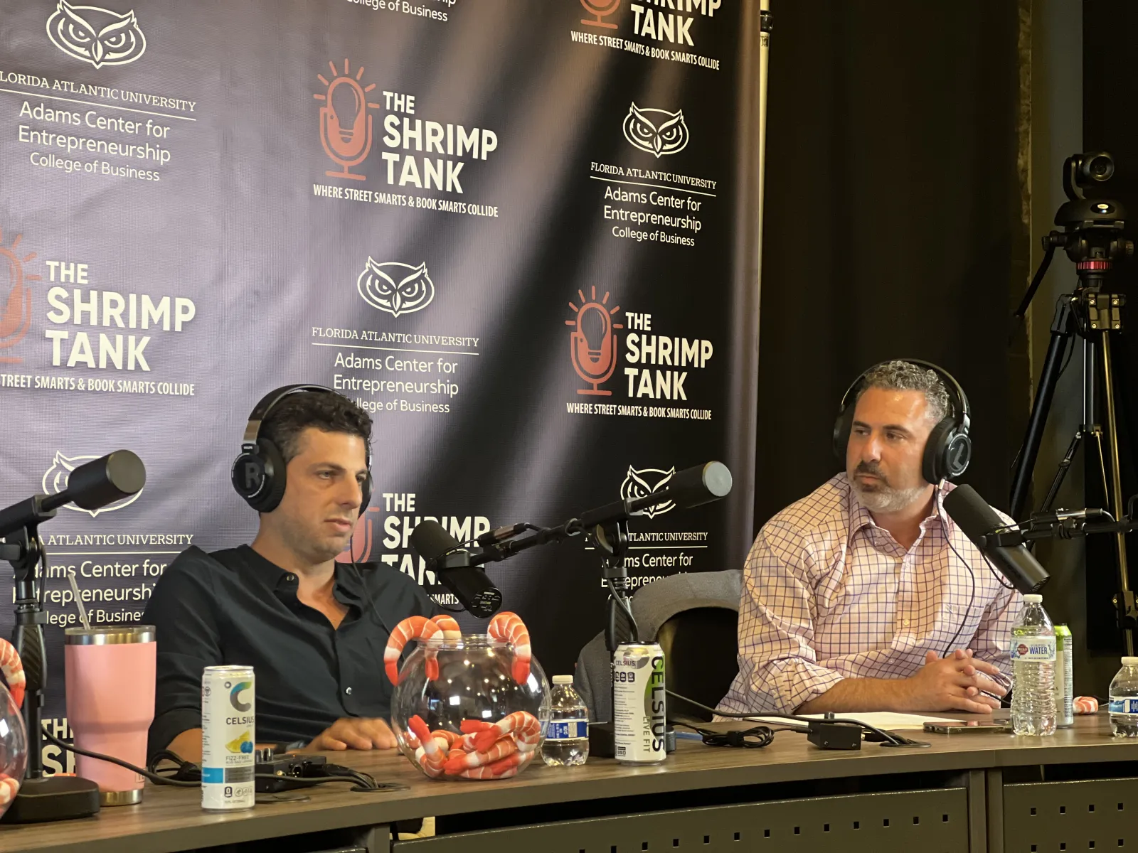 Two men podcasting on The Shrimp Tank with microphones, headphones, and branded backdrop in a studio setting.