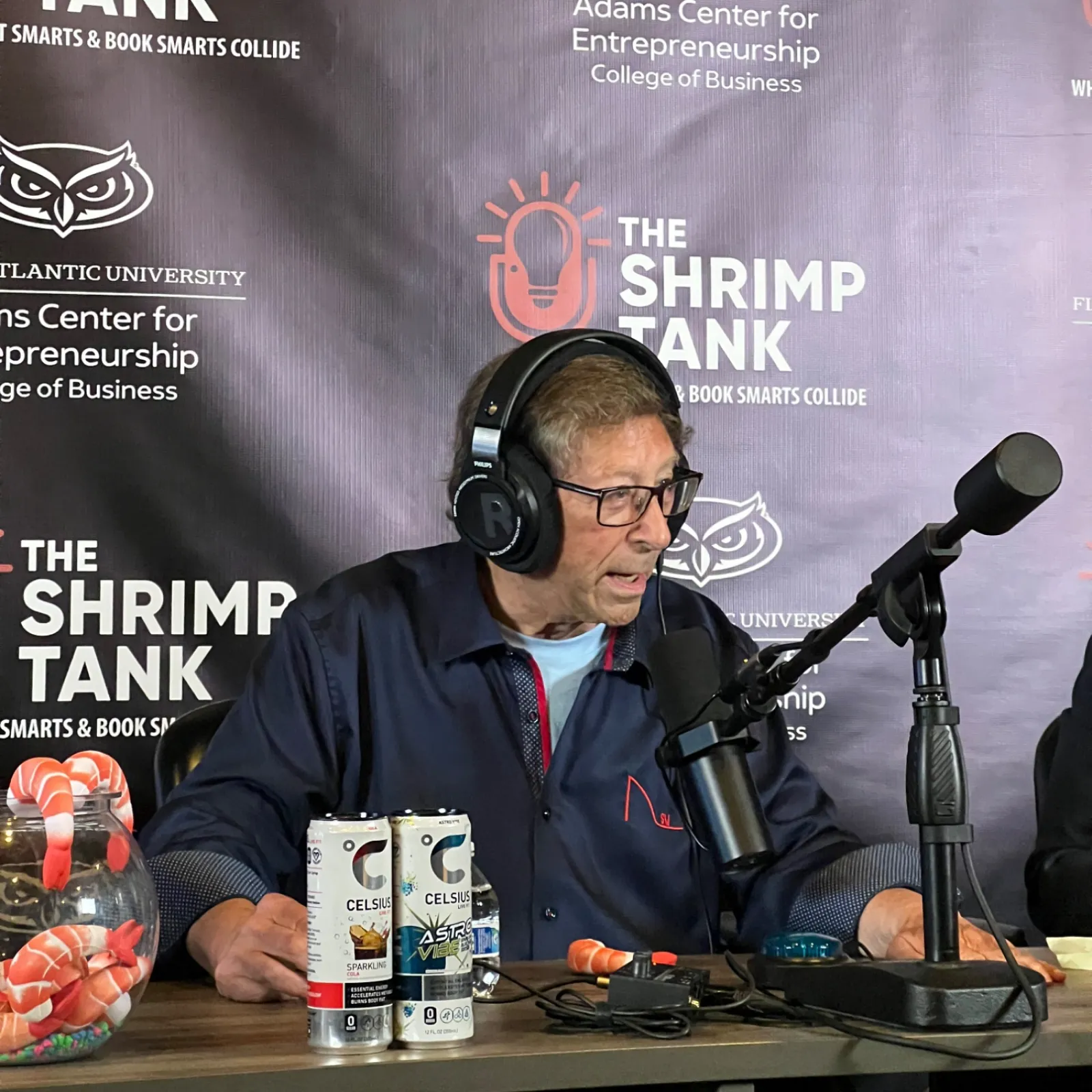 Man wearing headphones speaking into a microphone at The Shrimp Tank podcast with drink cans on the table