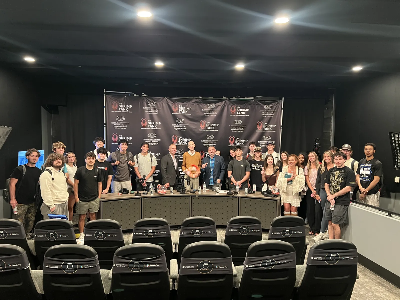 Group of people posing inside a studio with Shrimp Tank media banners and theater-style seating in the foreground