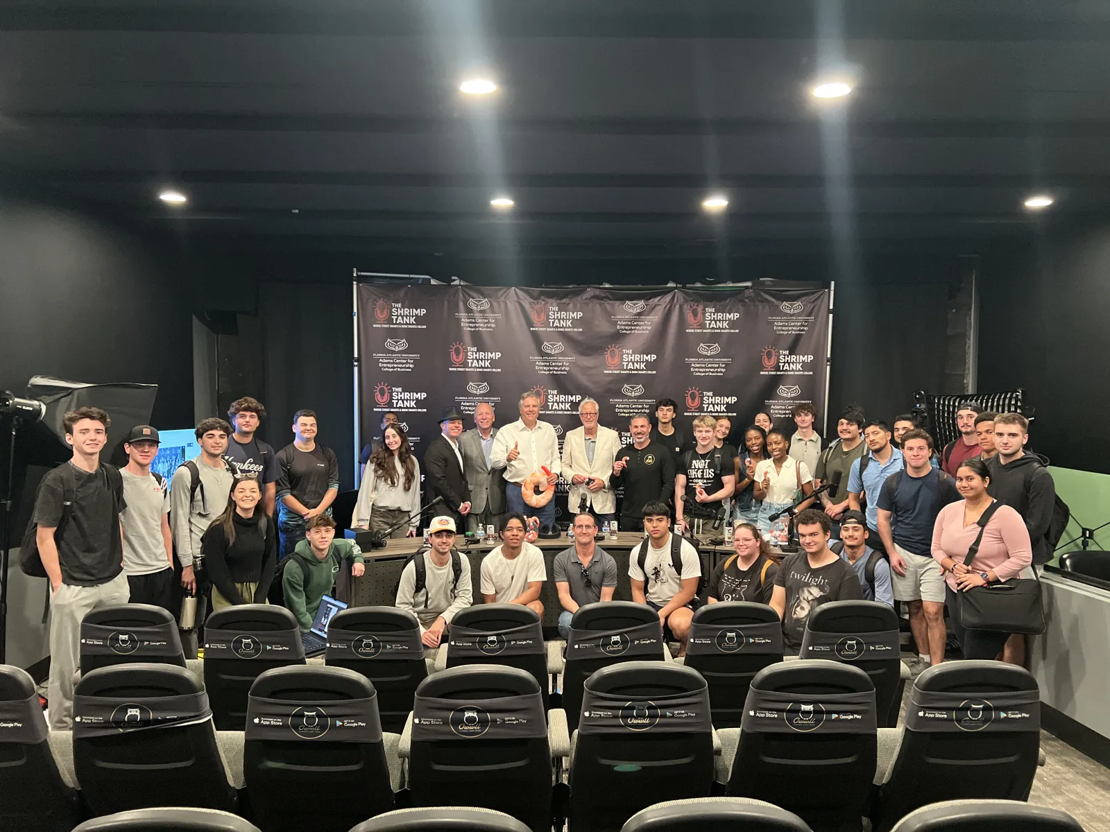 Large group of diverse young adults and speakers posing in front of a Shrimp Tank branded backdrop in auditorium.