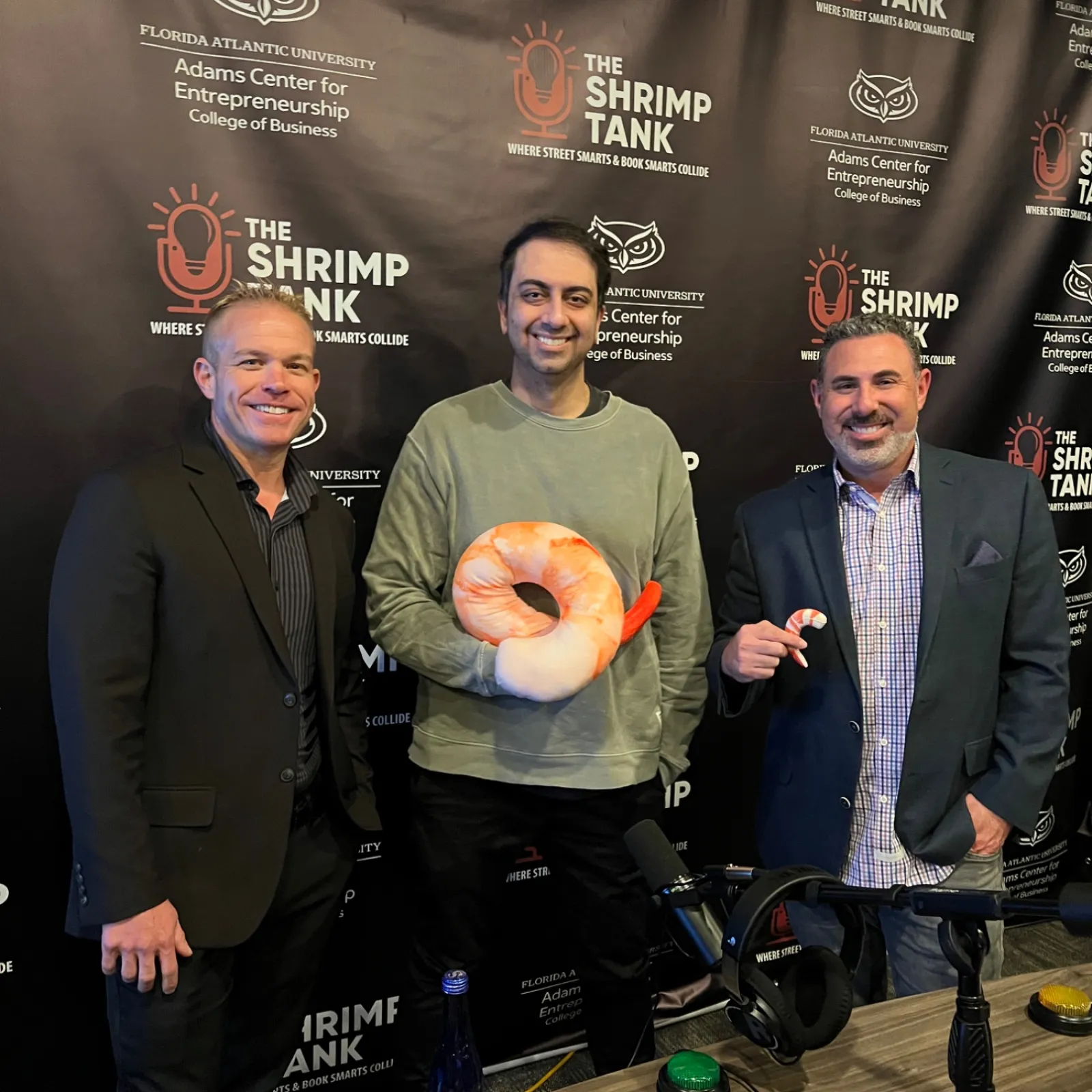 Three men posing at The Shrimp Tank event with shrimp plush toys and a branded backdrop of Florida Atlantic University.