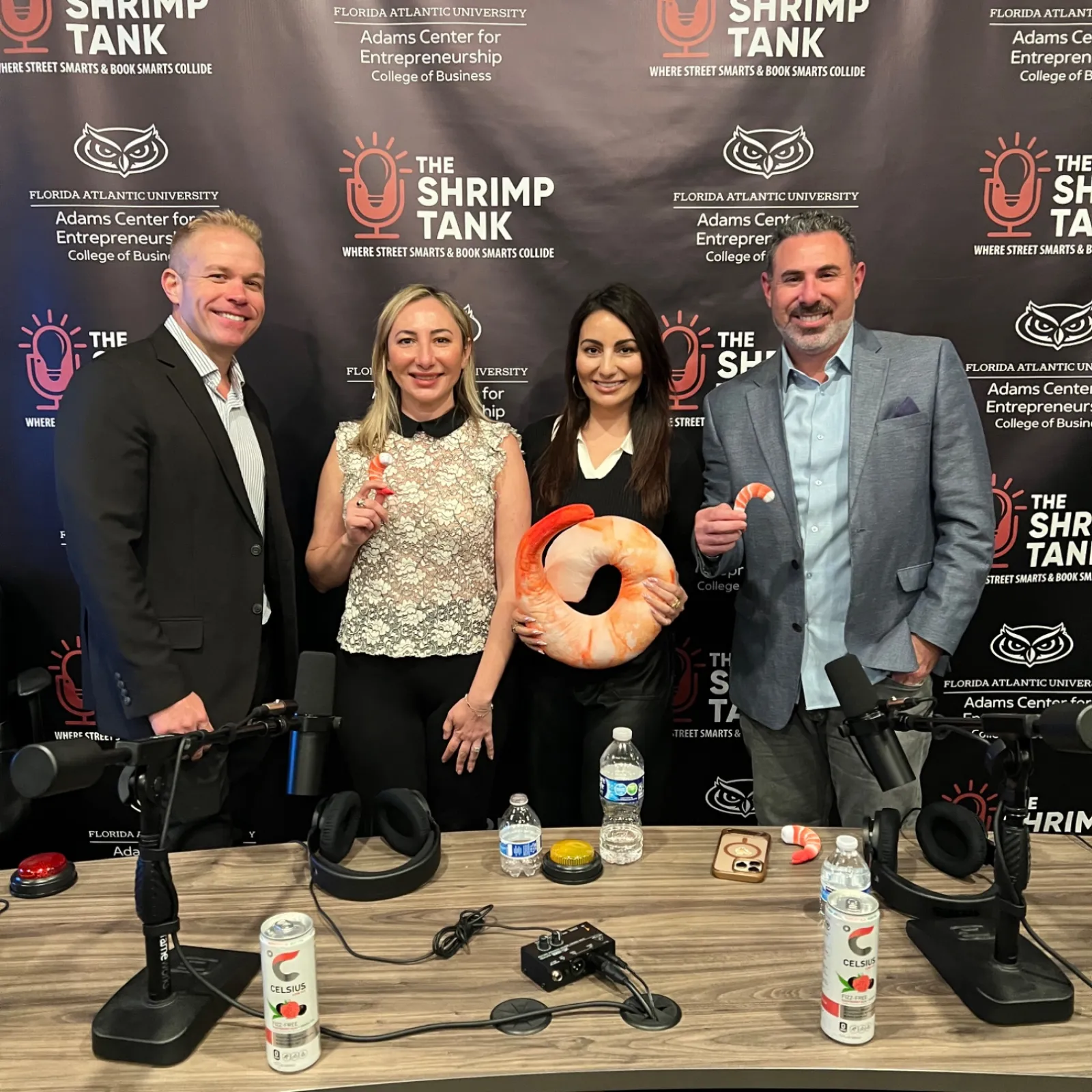 Four people posing with shrimp-themed items at a podcast studio with The Shrimp Tank backdrop.