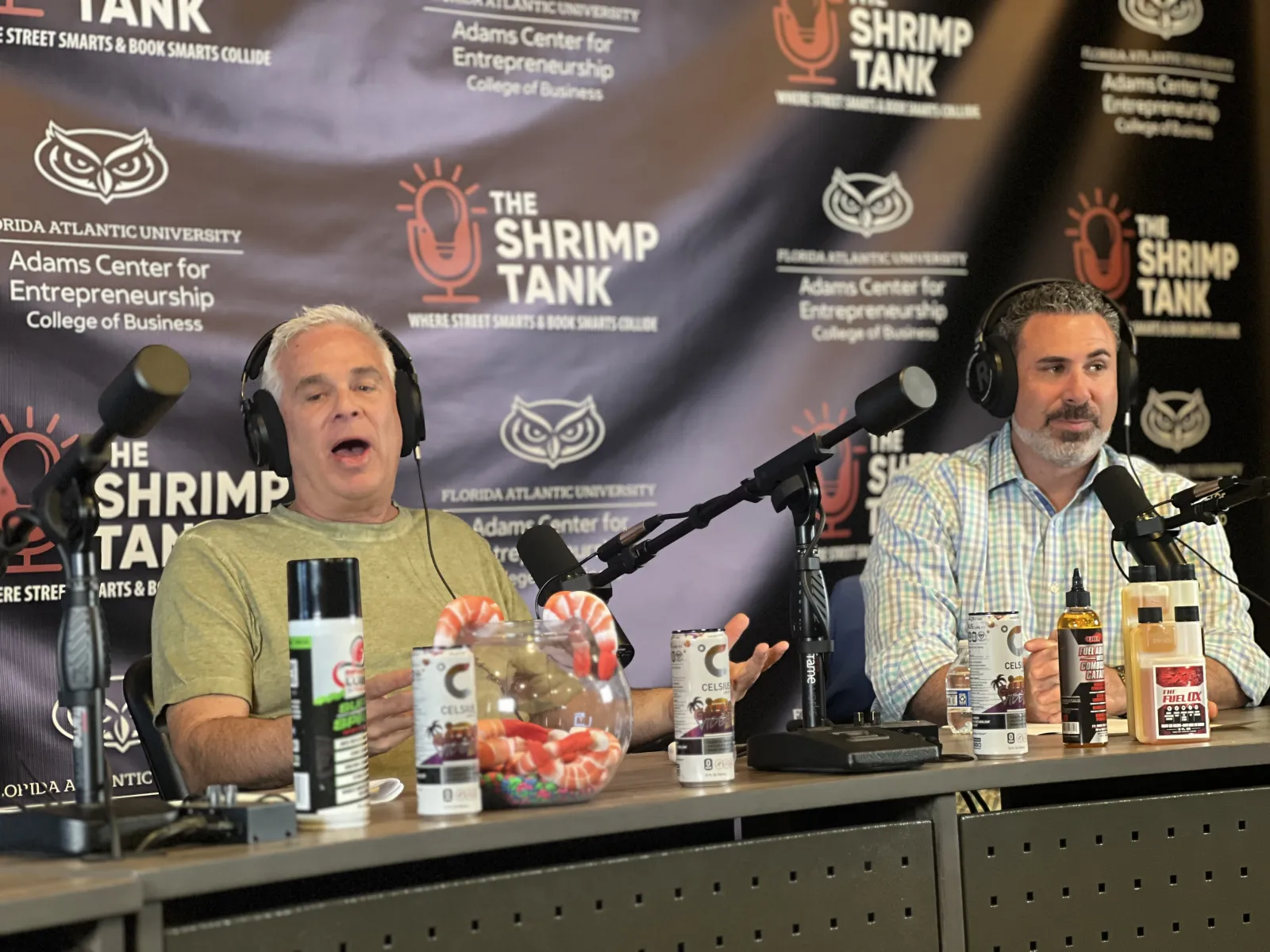 a couple of men sitting at a table with microphones and food