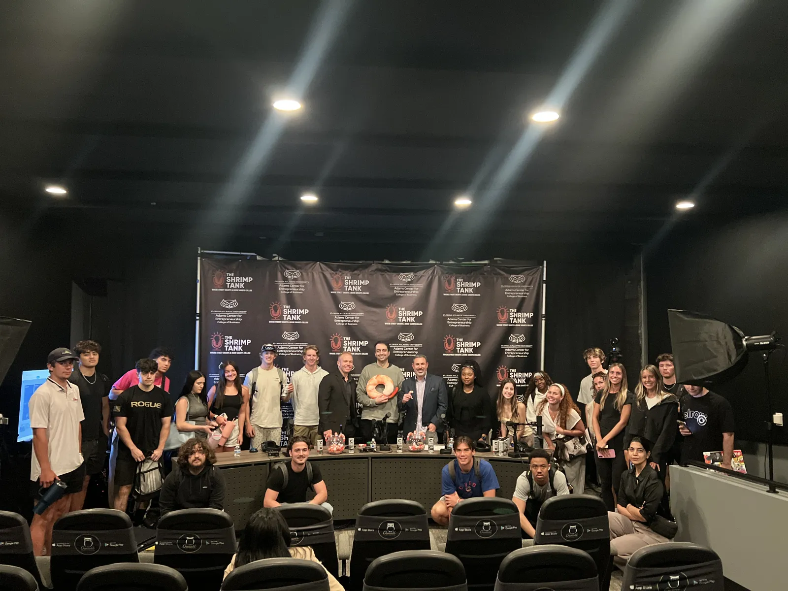Group of people posing in a studio with Shrimp Tank banners and professional lighting equipment in the background.
