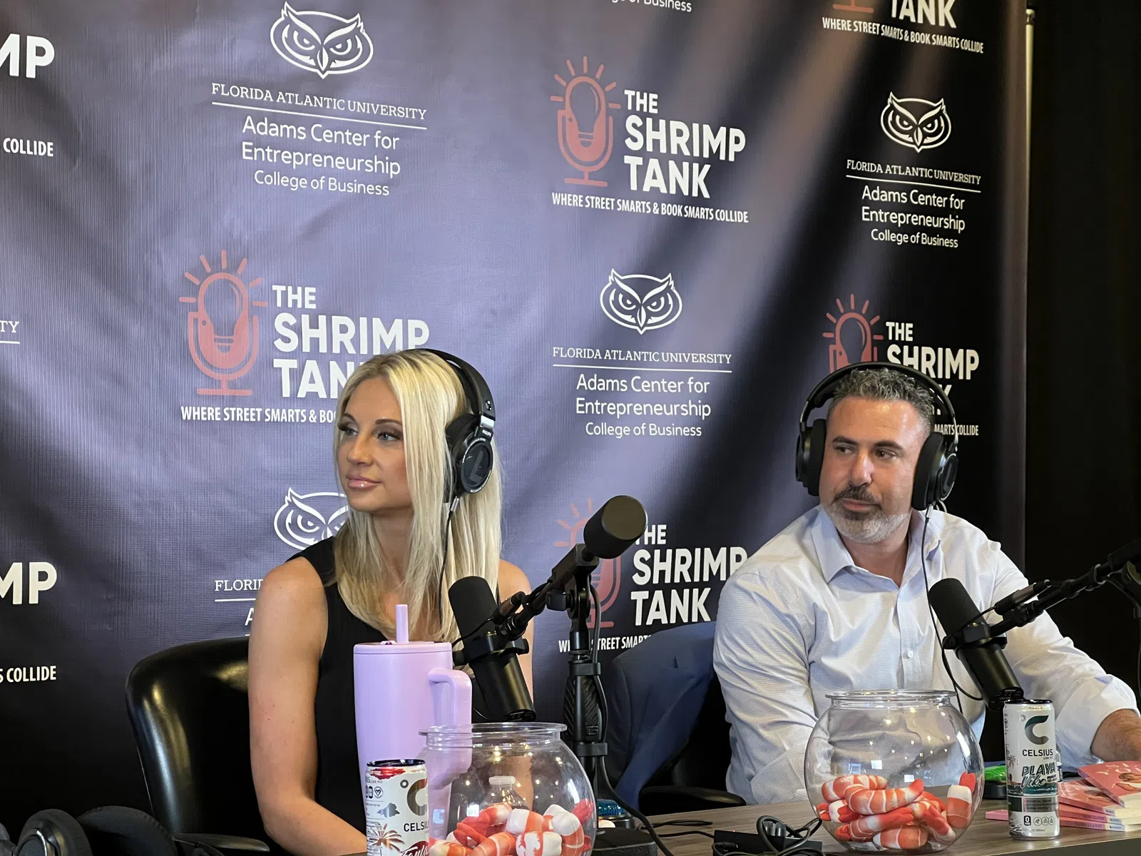 Two podcasters with headphones speaking into microphones at The Shrimp Tank podcast set with FAU branding.