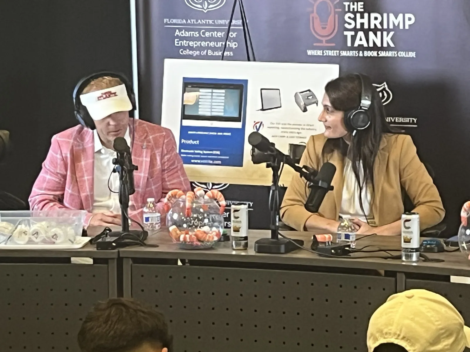 Two podcast hosts wearing headphones converse at a table with microphones and shrimp decor, Shrimp Tank event backdrop.